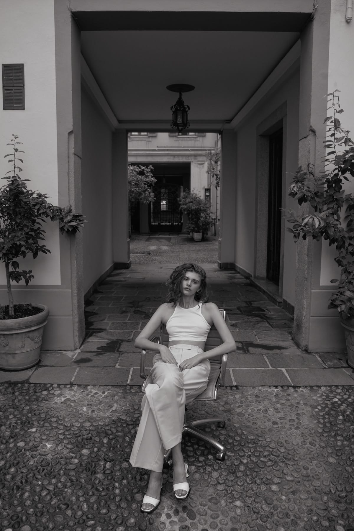 Alyona Lihosherstowa sits in an office chair set in a Milanese courtyard, in a white crop top, cream trousers and white sandals, photographed by Alexei Bazdarev.