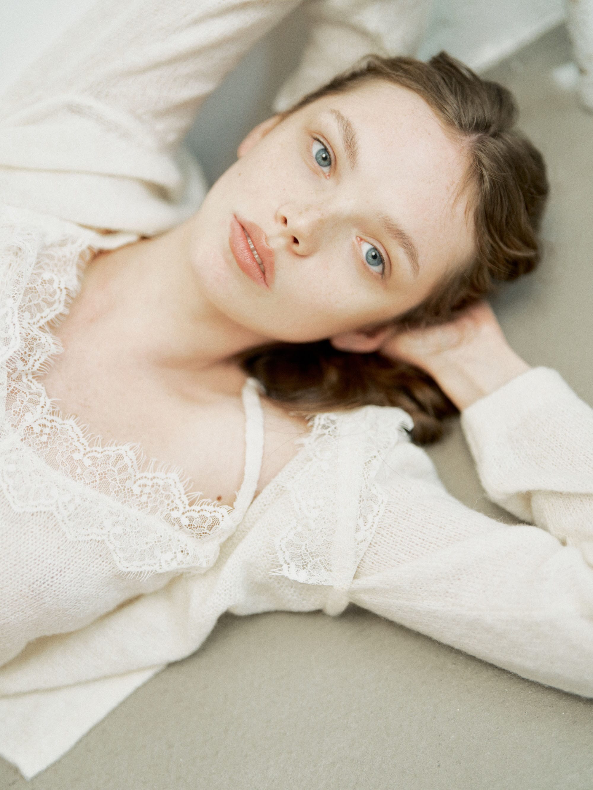Anna lies on her back looking up at the camera with hands tucked under her head, wearing a cream knit cardigan over a lace-trimmed camisole, photographed by Svetlana Latyshko.