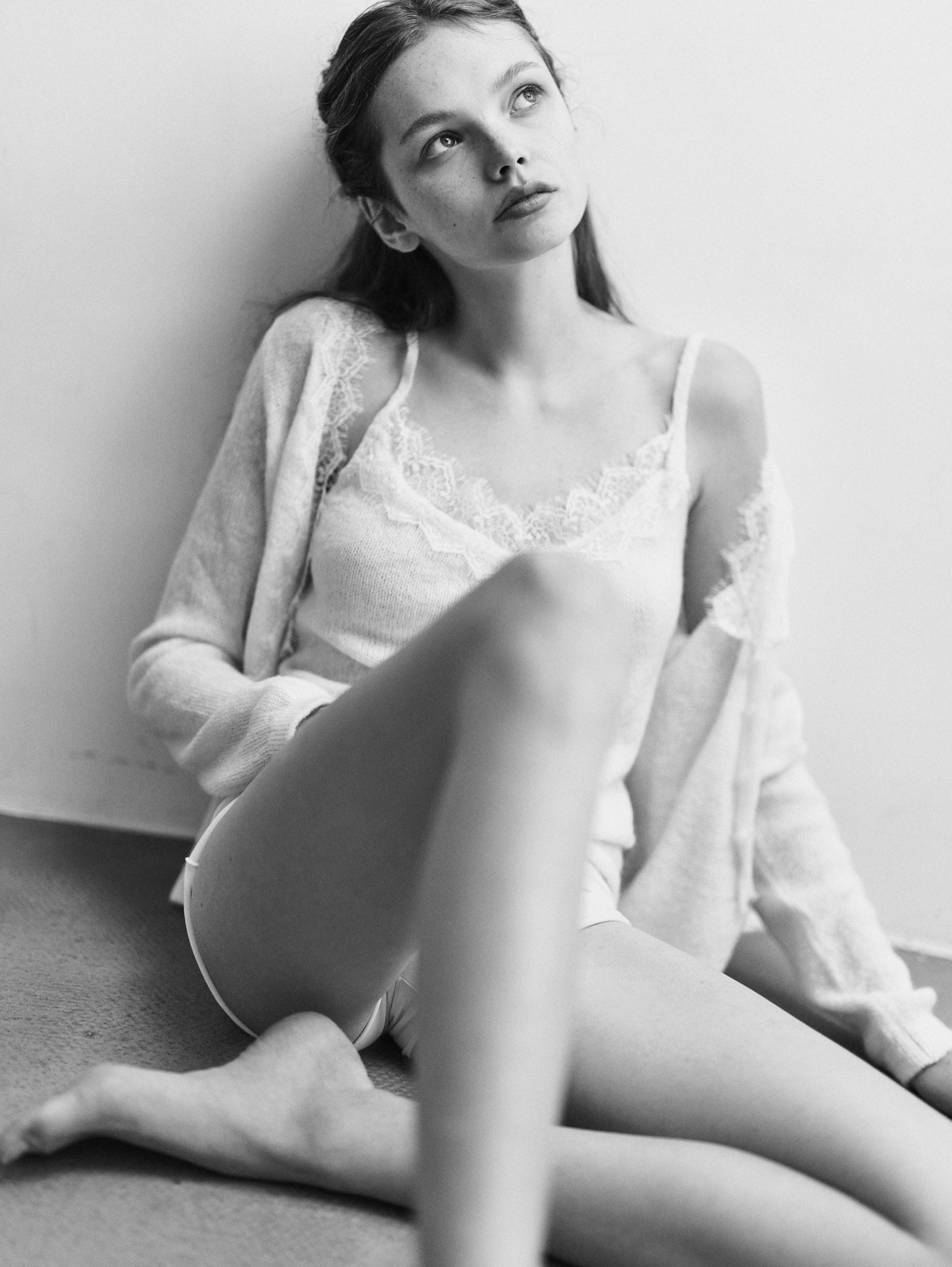 Anna sits against a wall with her knee drawn up in a lace-trimmed camisole, cardigan and white briefs, looking up and away from the camera, photographed by Svetlana Latyshko.