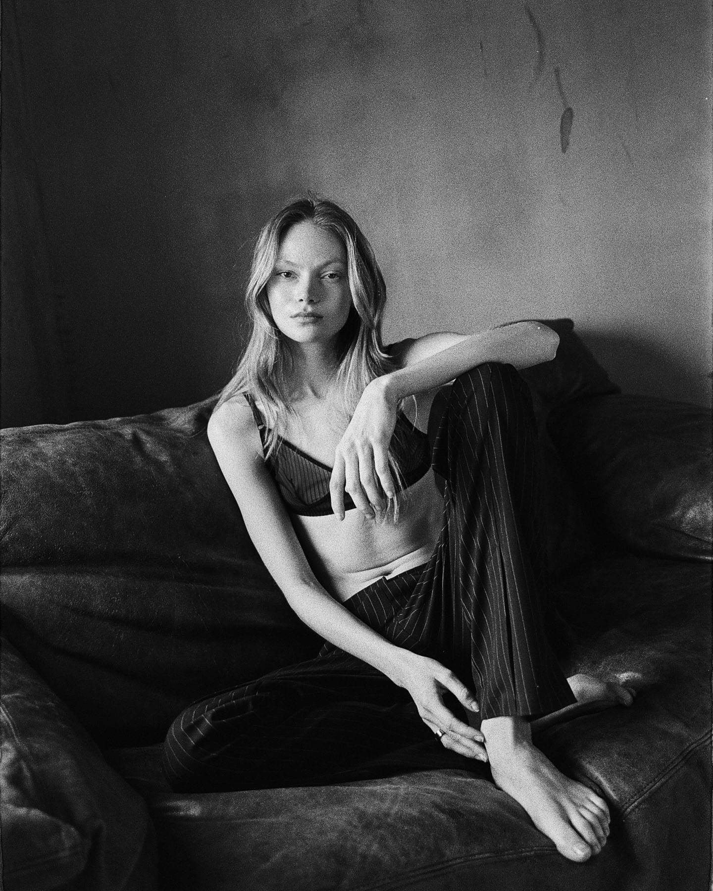 Maria Privalova sits on a leather sofa in a pinstripe bralette and matching trousers, barefoot, meeting the camera, by Natasha Skatnikova.