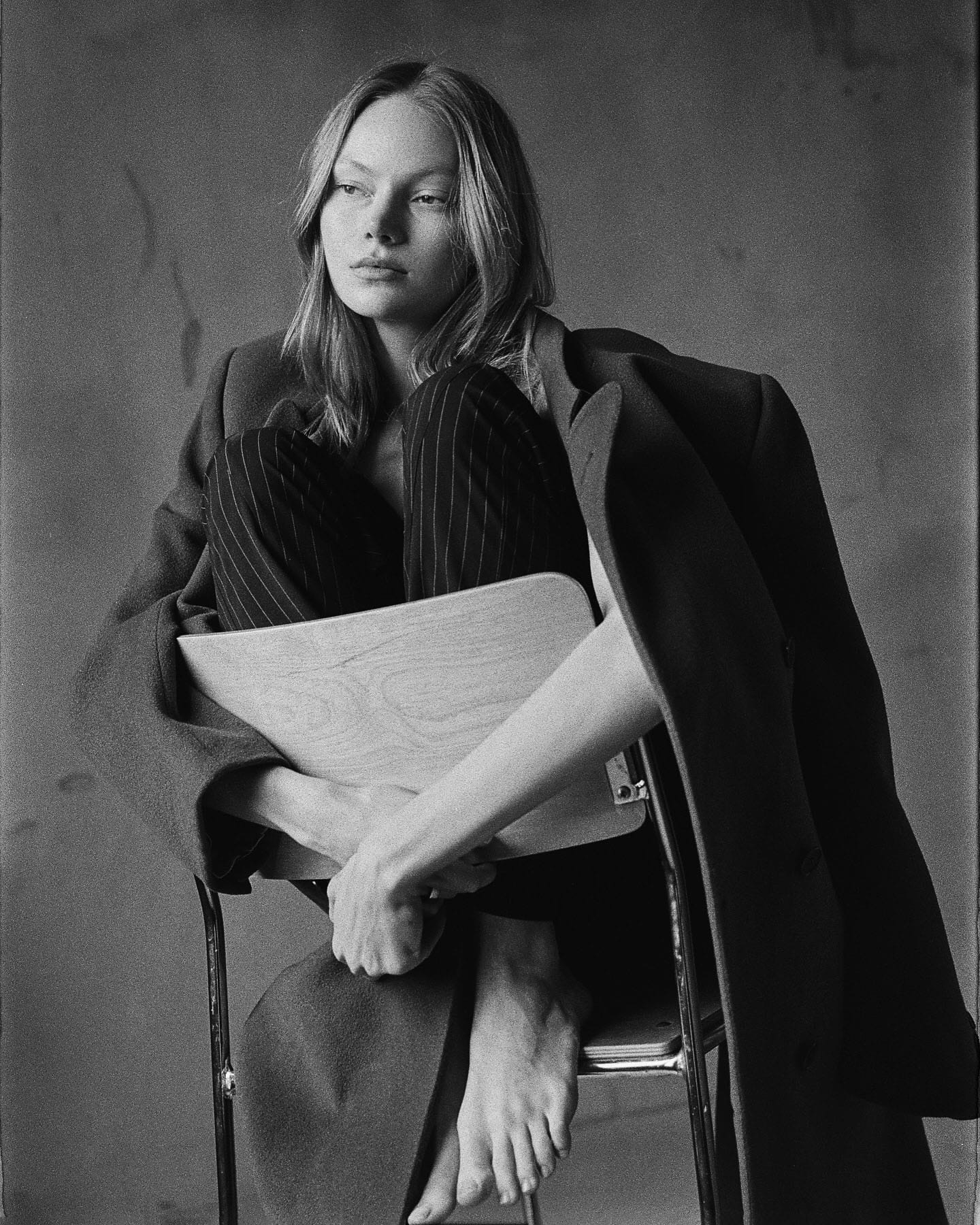 Maria Privalova sits on a chair, knees drawn to her chest under an oversized coat, looking away, by Natasha Skatnikova.