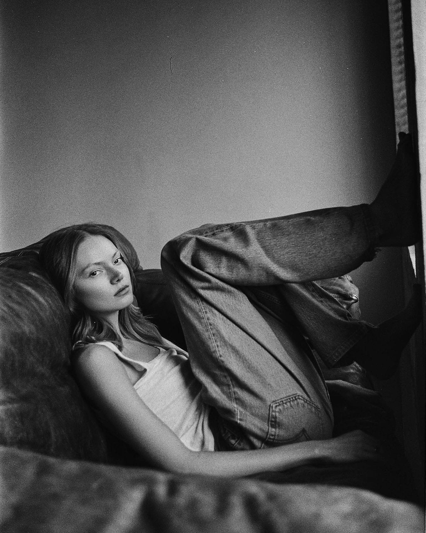 Maria Privalova reclines in a leather sofa in a white tank and jeans, legs up the wall, looking into the lens, by Natasha Skatnikova.