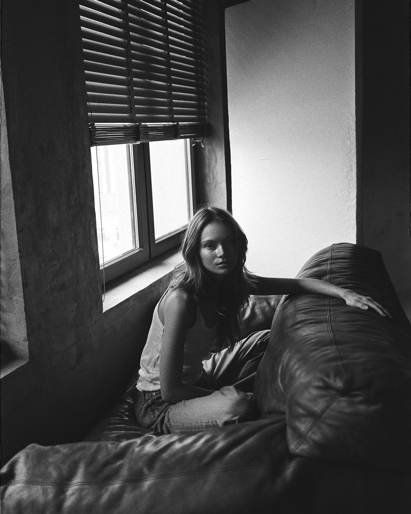Maria Privalova sits in a window nook by raised blinds, arm along a leather sofa, in a tank and jeans, by Natasha Skatnikova.