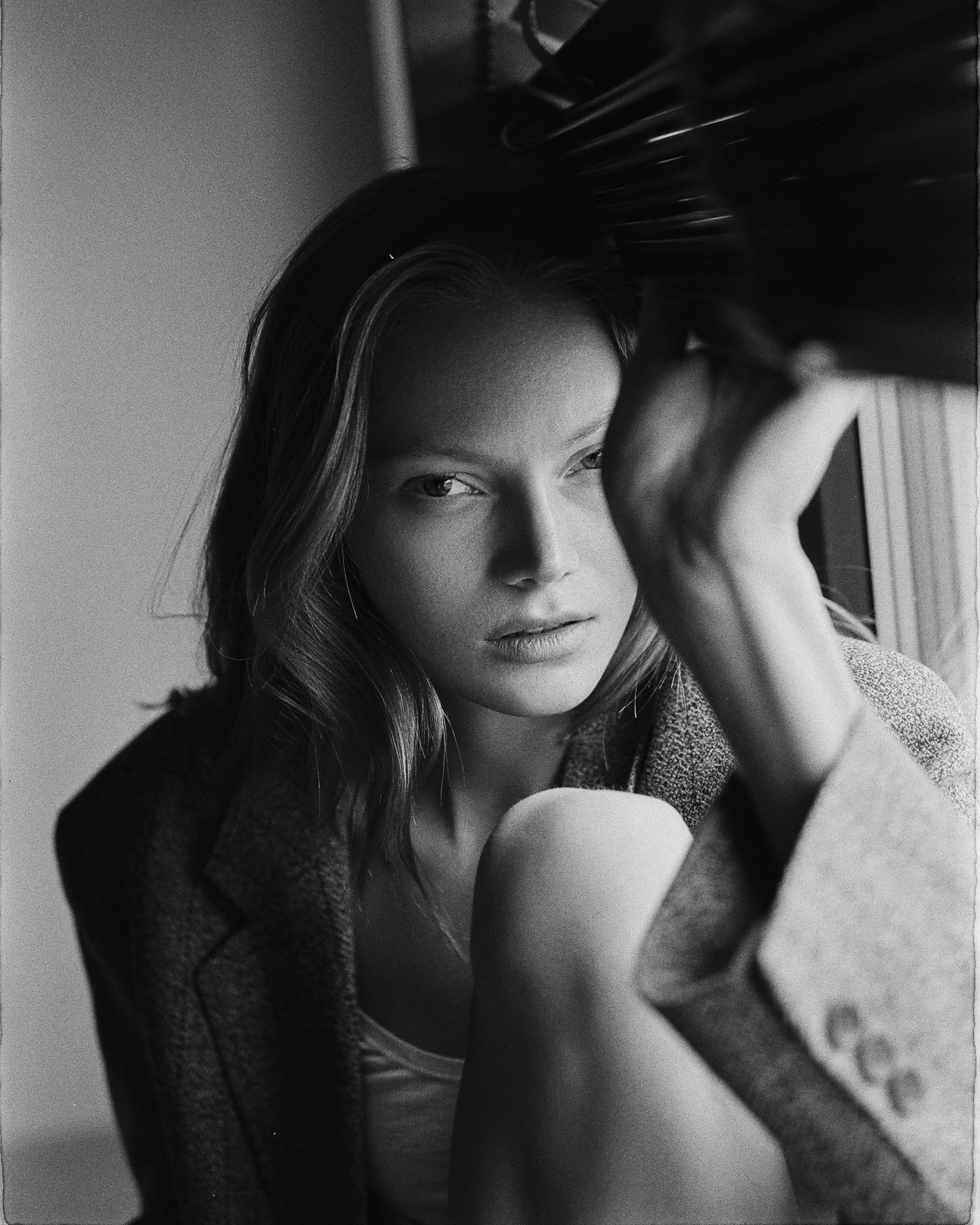 Maria Privalova lifts a window blind, knee raised, in a tweed jacket over a light top, gazing at the camera, by Natasha Skatnikova.