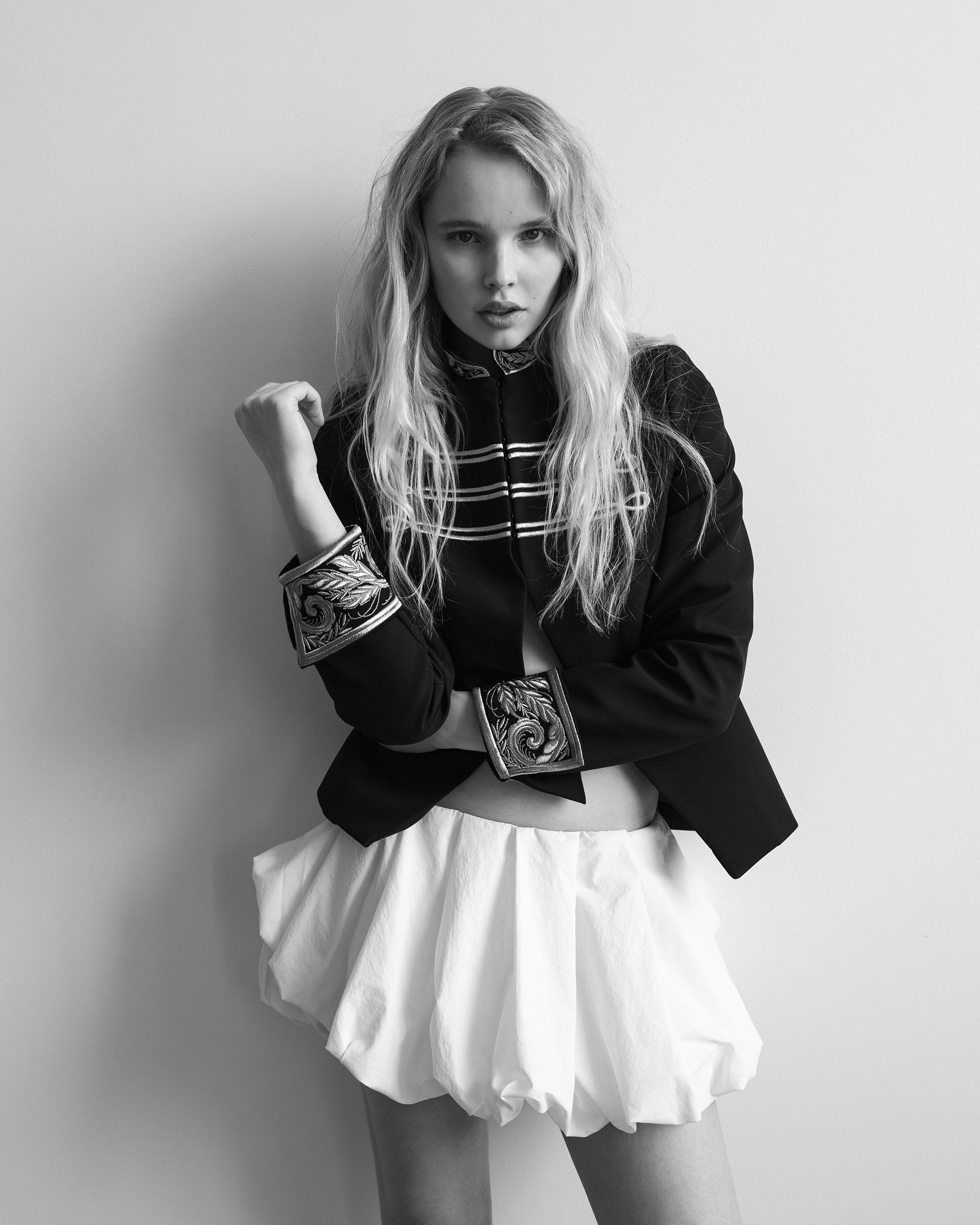 Jeanne Markevich in a black military jacket and white bubble mini, hands at her chest, by Kristina Ranneva.