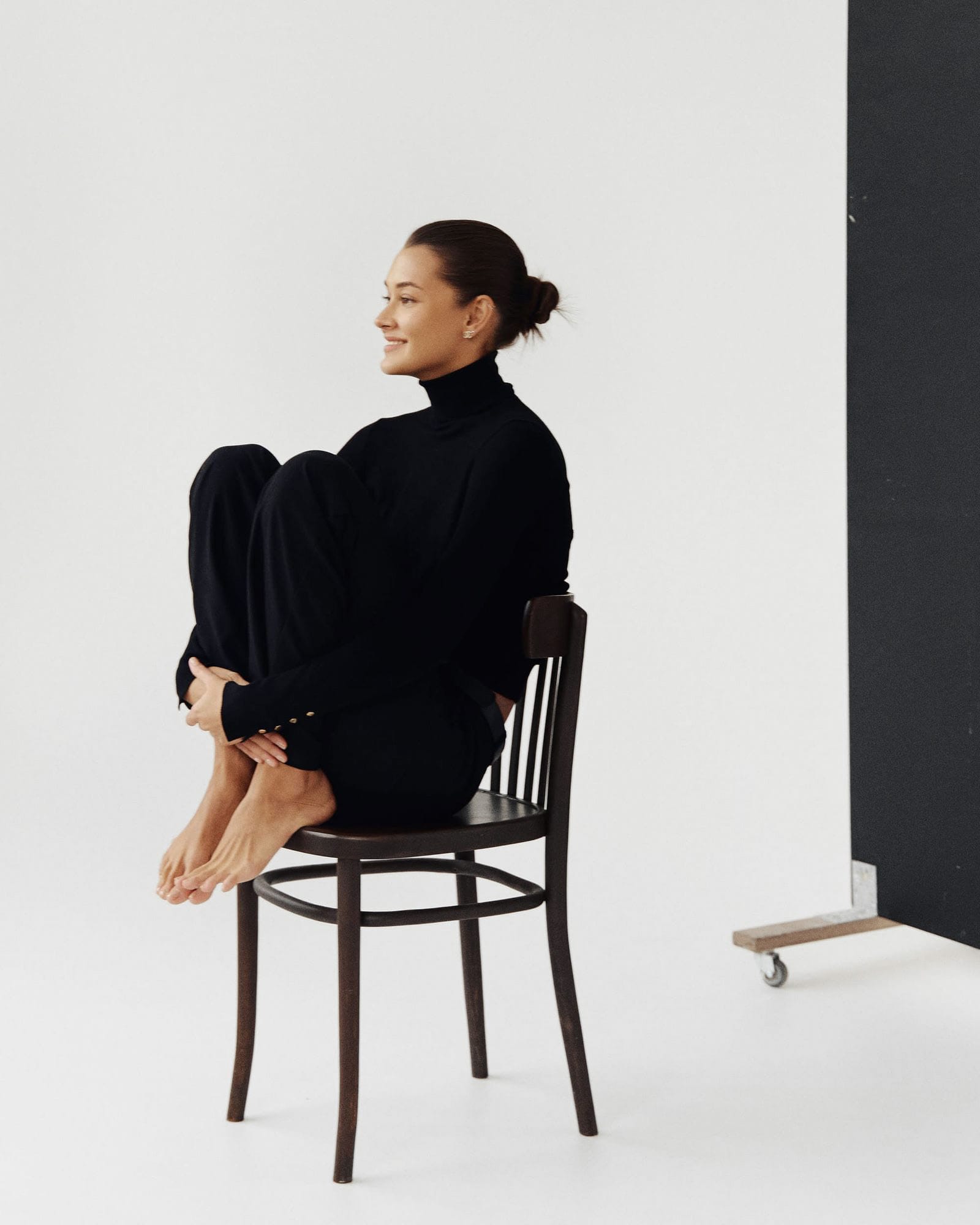 Kristina Yakovleva sits on a bentwood chair against a white backdrop, smiling in profile, in all black, by Maxim Kabanov.