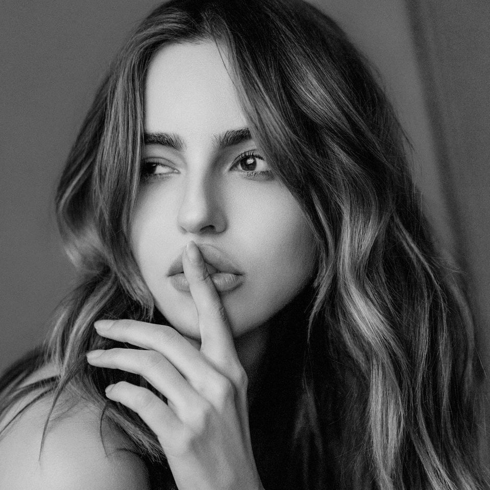 Black-and-white close-up of a young woman with long, wavy hair, one finger pressed to her lips, looking away from the camera.