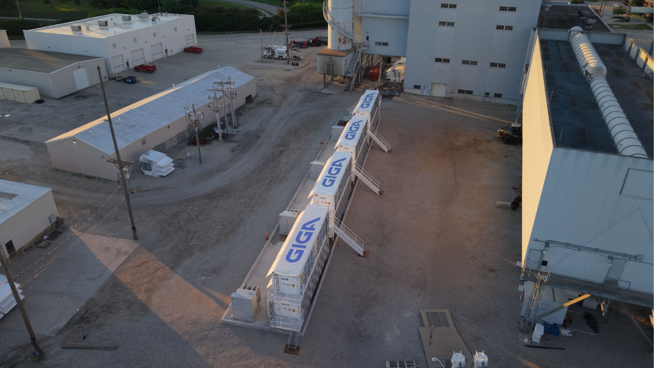 An aerial view of the Simple Mining facility in Iowa featuring multiple white GIGA containers arranged in a row next to industrial buildings during sunset.