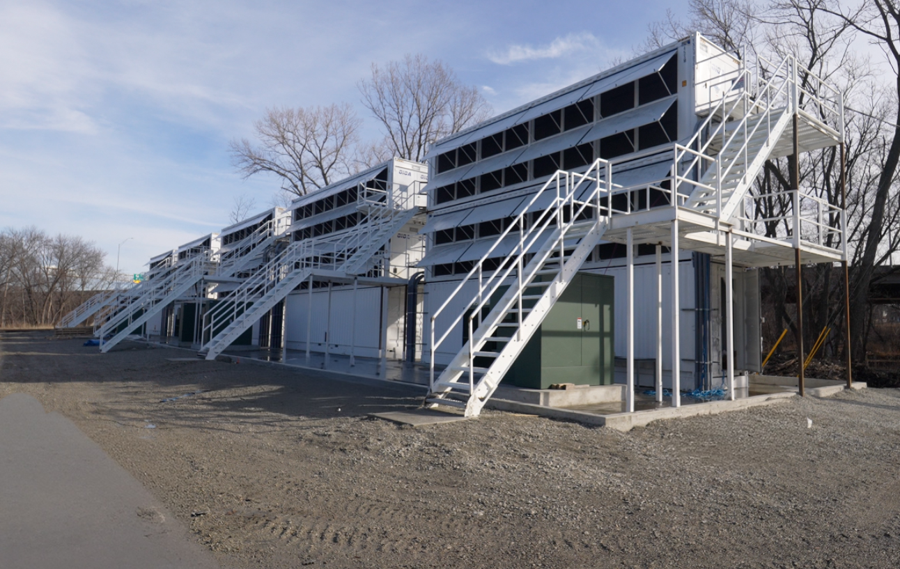 Row of white industrial Bitcoin mining containers in an outdoor gravel lot, featuring exterior louvers for cooling and metal stairs for technician access.