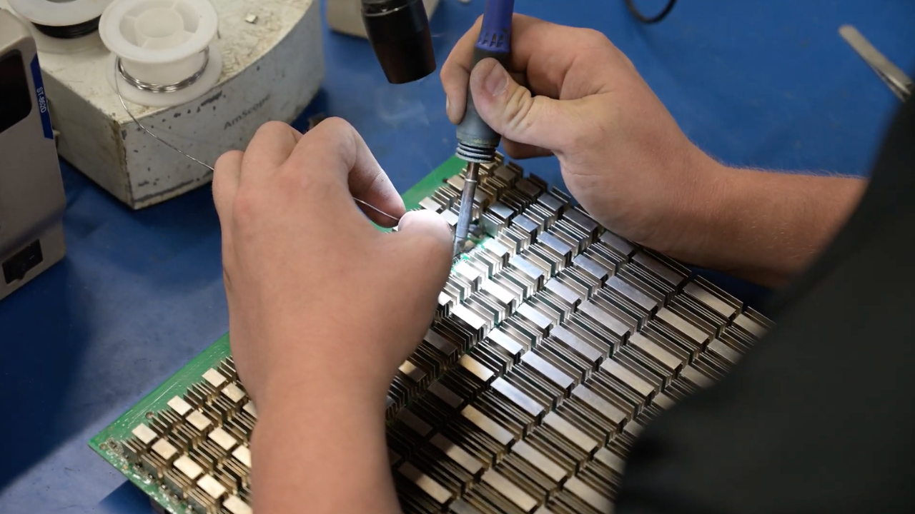 Close-up photograph of a technician using a soldering iron and wire to perform precise maintenance on a dense ASIC miner hashboard covered in dozens of identical aluminum heatsinks.