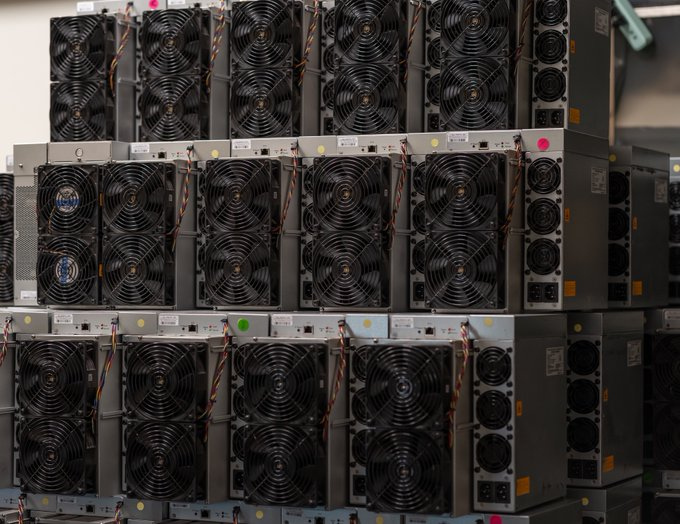 Rows and stacks of used Bitmain Antminer Bitcoin miners at a Simple Mining facility, showcasing the industrial metallic chassis and organized storage of secondary market hardware.