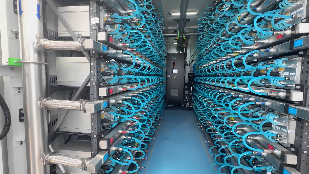 Aisle view inside a Simple Mining hosting facility showing two parallel rows of hydro-cooled Bitcoin ASIC miners. Blue liquid-cooling tubes connect each unit to a shared coolant loop, illustrating the kind of hosted infrastructure that runs Antminer S21 XP Hydro and S23 Hydro hardware at scale.