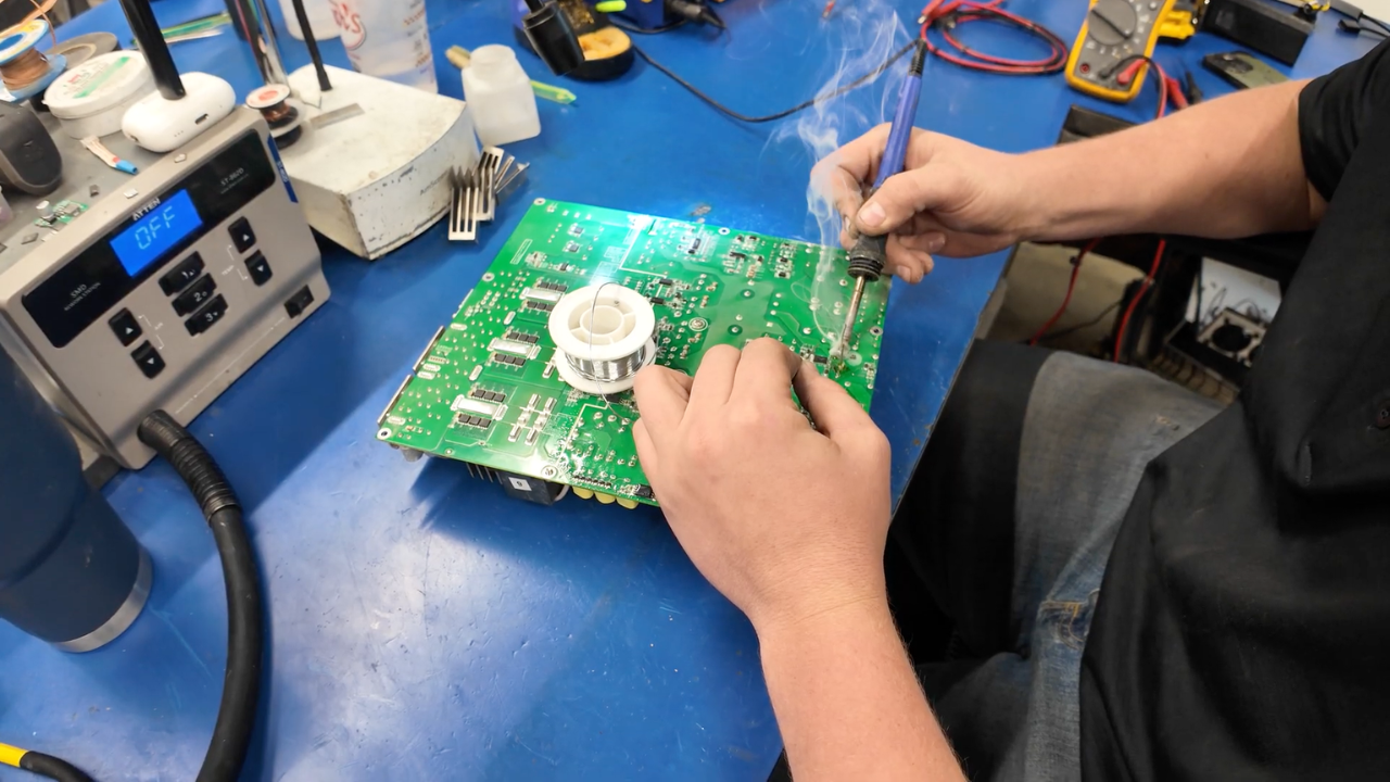 A Simple Mining technician uses a soldering iron to repair a Bitcoin ASIC miner board at the Iowa repair bench, with diagnostic tools alongside.