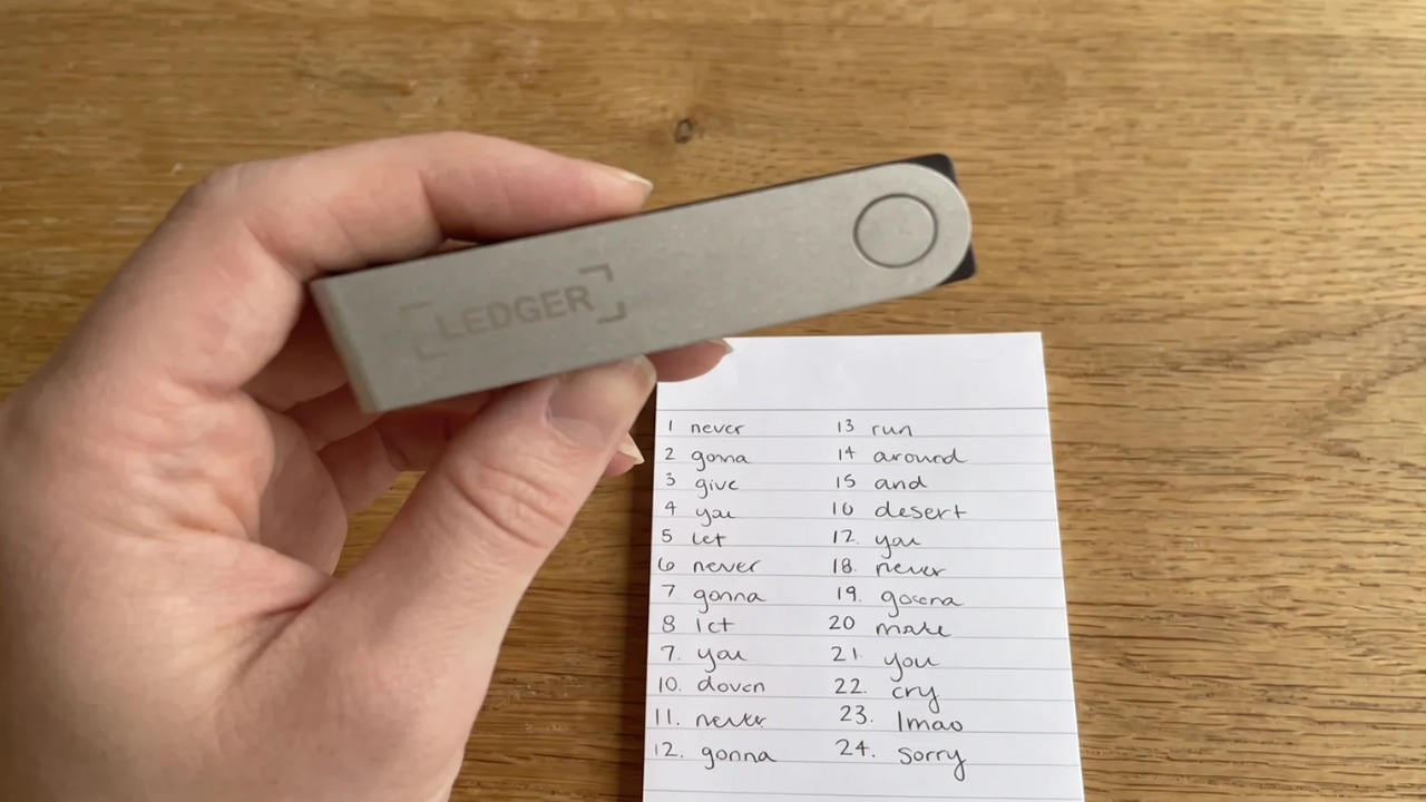 A hand holding a Ledger hardware wallet beside a handwritten 24-word seed phrase list on paper, showing a Bitcoin cold-storage backup setup.