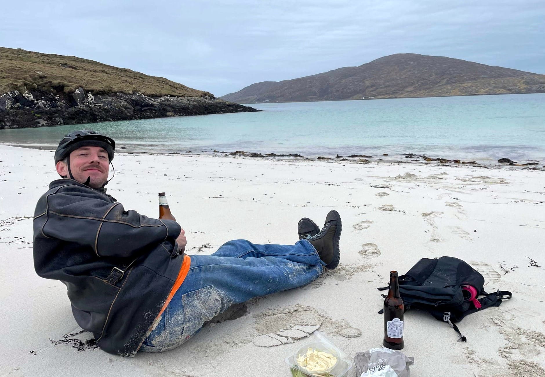 A man is sitting smiling on a white sand beach in a bay with blue sea water. He has a beer in his handle, a bike helmet on his head, and there's a small hill in the background.