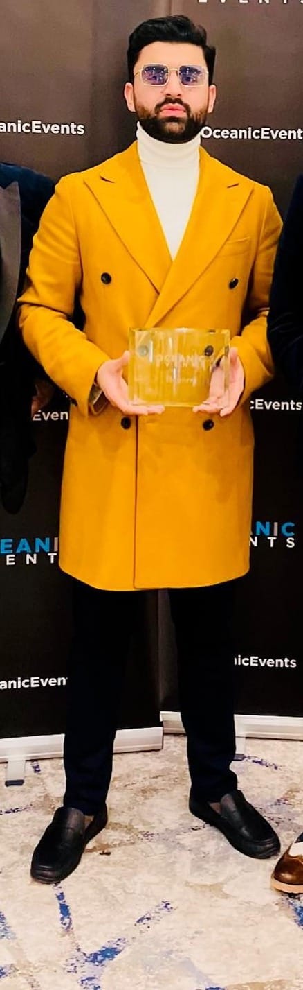 A man stands in a bright yellow smart jacket in blue sunglasses holding an award he won.