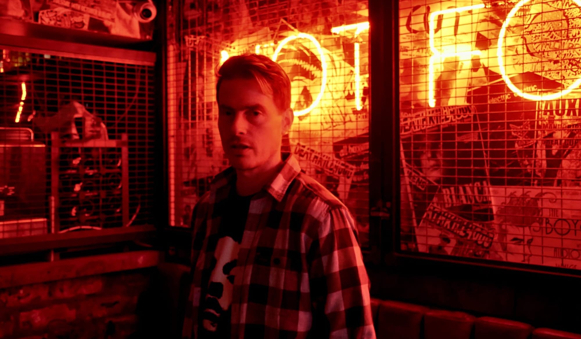 A man looks at the camera. his face is lit red by a large red neon sign behind him. He has short red hair which is swept to one side. 