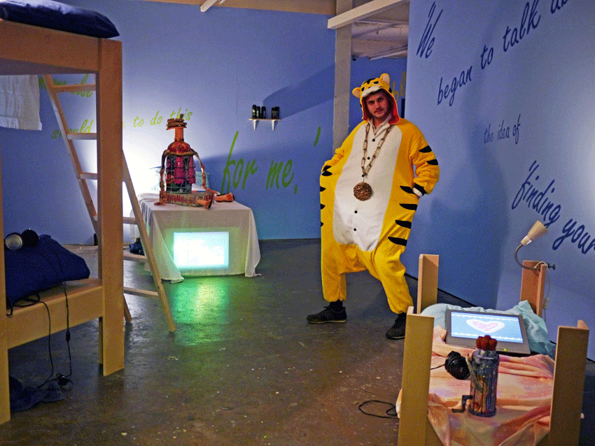 Art installation with a man posing in a tiger onesie in a bedroom with a bunkbed and purple walls.