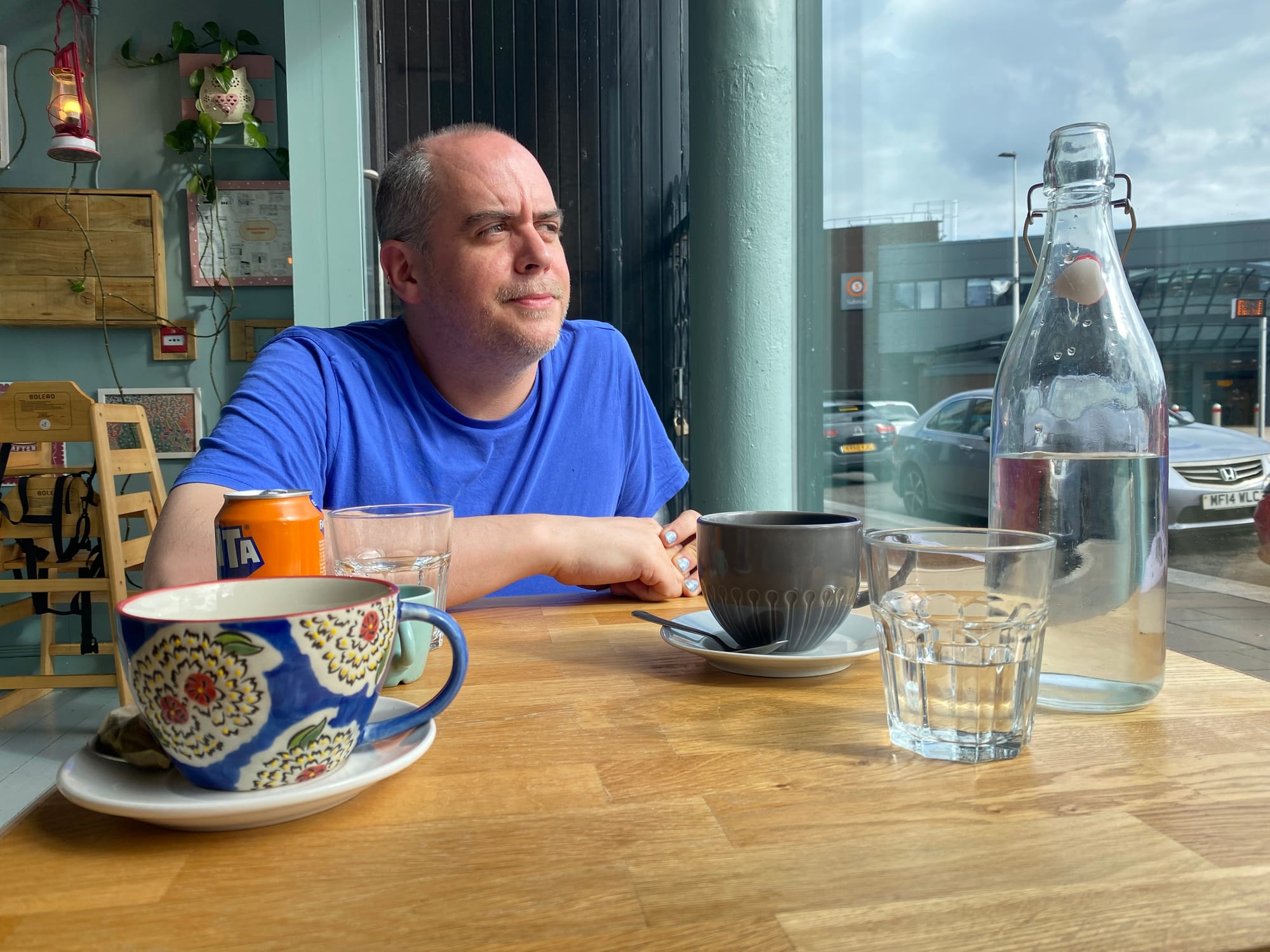 Man in a blue tee shirt with balding short hair looks out the window of a cafe. 