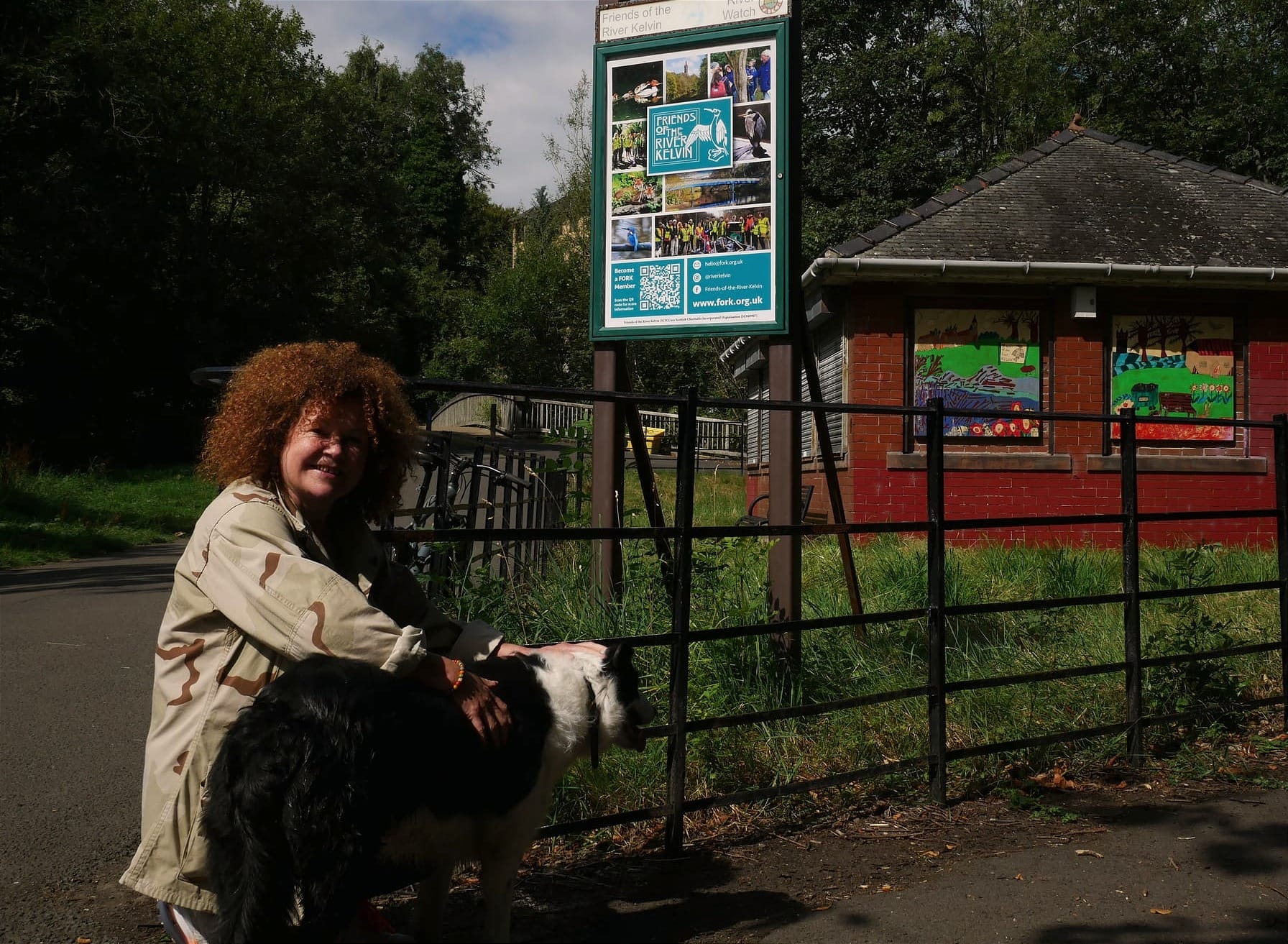 A woman with red frizzy hair kneels smiling next to a border collie. She is outside a small red brick building.