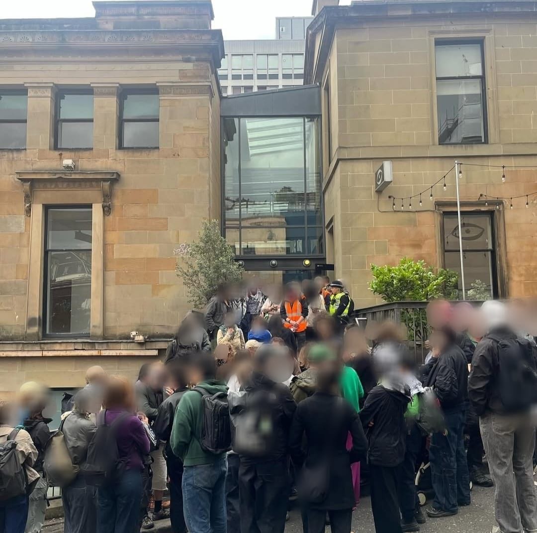 A group of protestors with blurred-out faces stand on Scott Street in Glasgow. Some are on the steps of the side entrance to the CCA, as well as police. 