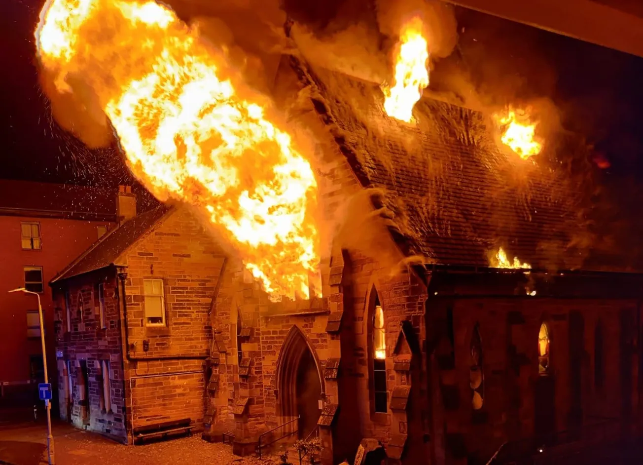 A dramatic image of a huge ball of flames emerging from a church window. There are also flames licking up through smaller windows on the roof. 