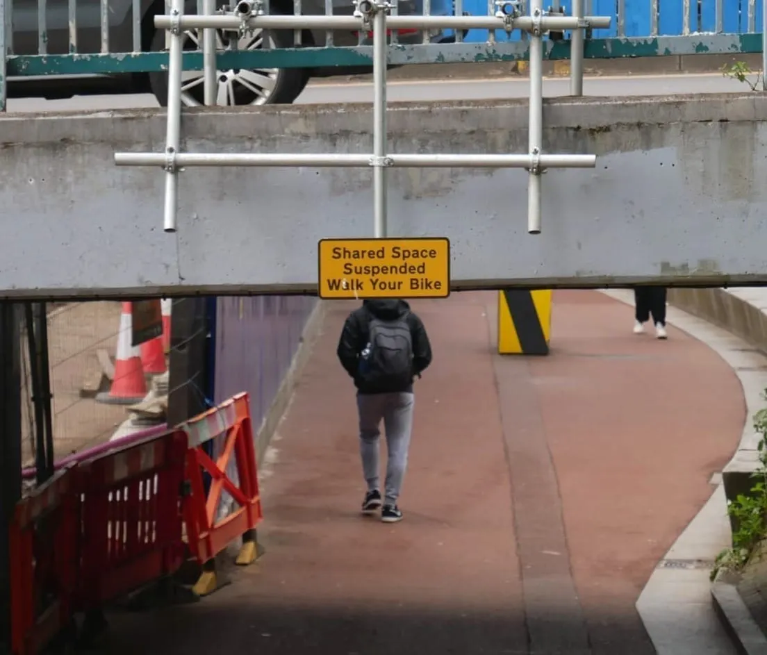 A man with a rucksack on walks away from the camera through an underpass. His head isn't visible, in front of it is a road sign.