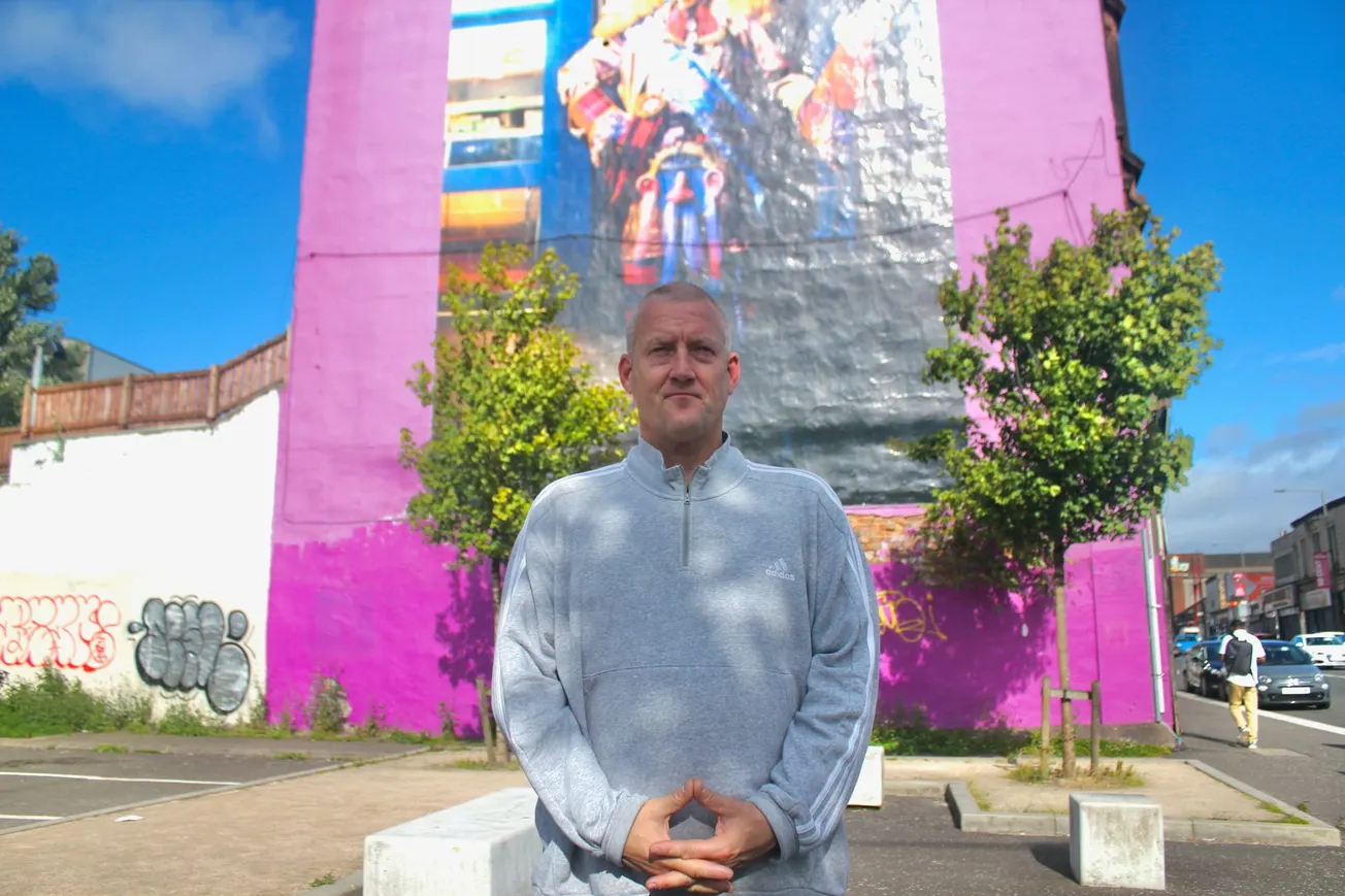 A man with a shaved head in a grey pullover stands in front of a bright pink mural. 