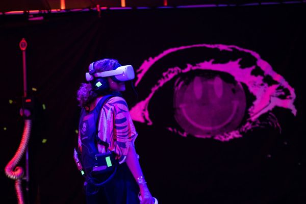 En kvinna i VR glasögon framför ett stort målat öga i neonrosa. 