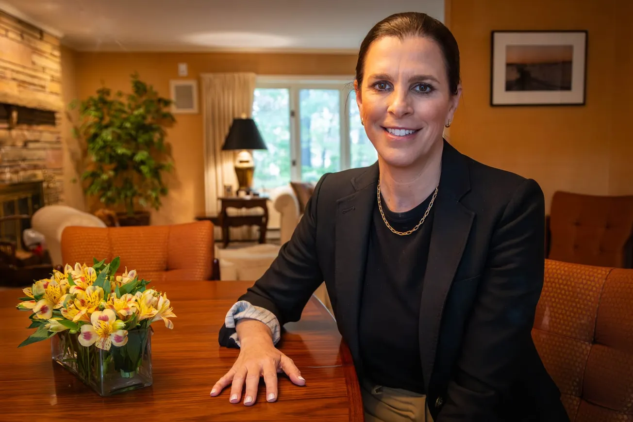 Woman in dark blazer seated at a wooden table with flowers in a warmly lit living room.