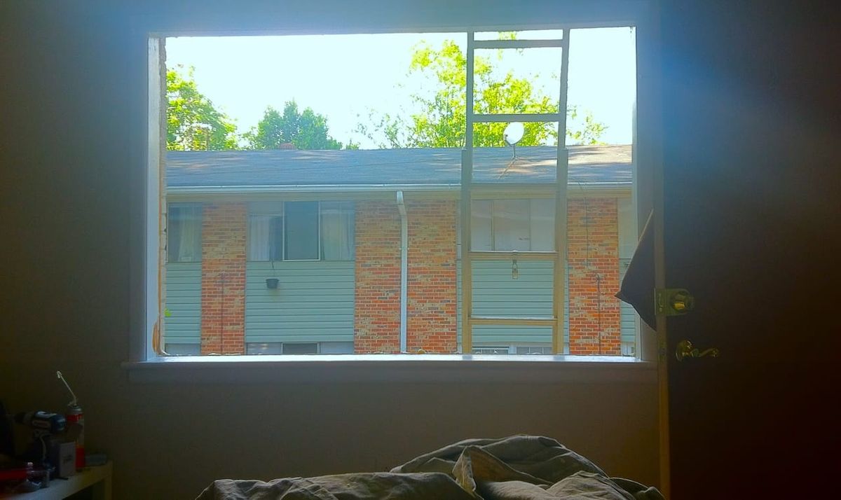 a hole in an exterior wall where a window should be. an open door frames one side of the room while a small table with a bunch of stuff sits on the left.