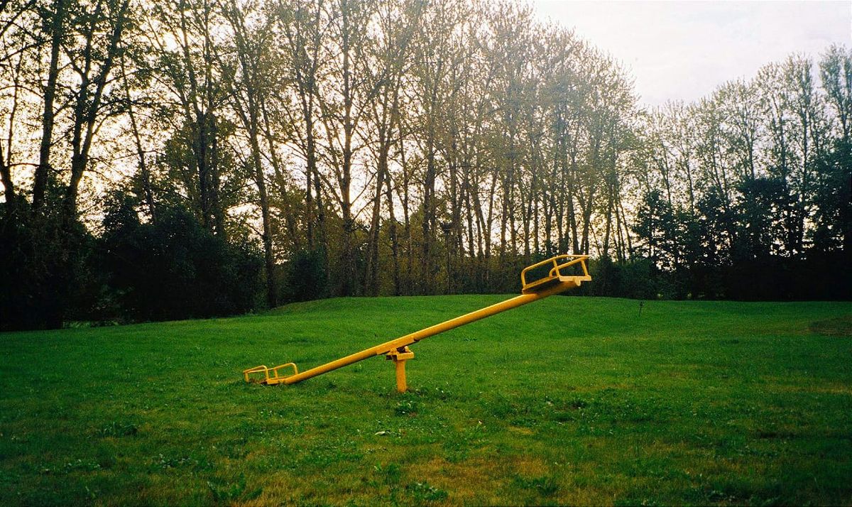 a yellow seesaw planted in the middle of a grassy field. thin trees line the periphery. their leaves and branches practically fade into the bright sky.