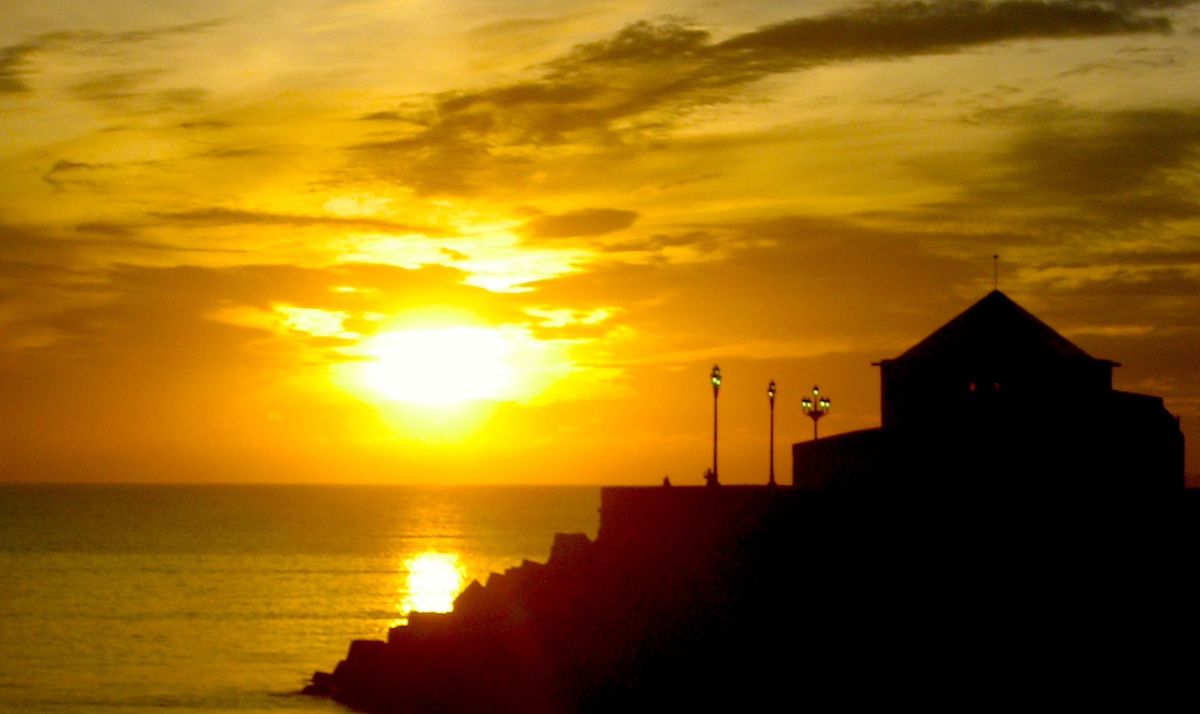 a photo of a sunset on the rocky shores of Cádiz, Spain. the still-bright yellow sun tints the sky golden. a building with a