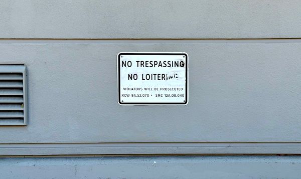 a sign against a gray building wall that reads NO TRESPASSING NO LOITERING with a threat of prosecution.