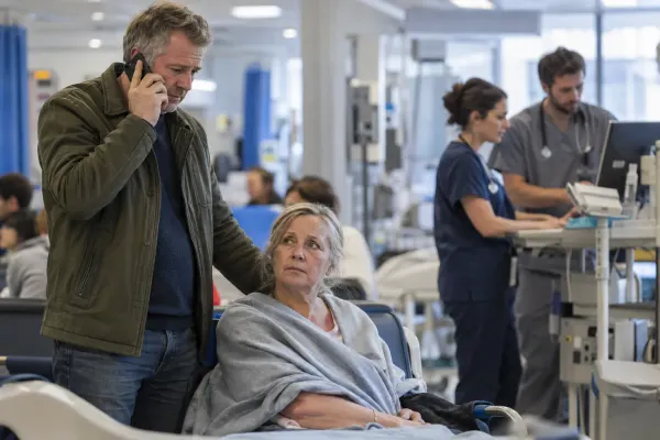 A family member calls for help beside a patient in a busy hospital emergency department while clinicians review care in the background.