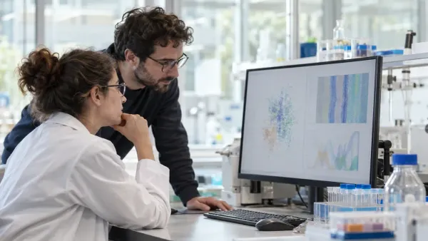 A laboratory scientist and a computational researcher review abstract data visualizations at an unbranded pharmaceutical research workstation.