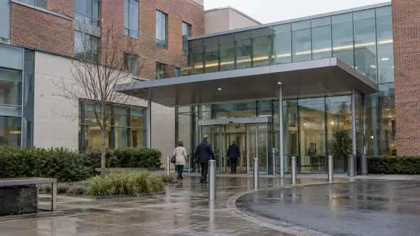 A modern unbranded British hospital entrance on an overcast morning with three anonymous adults walking toward the doors
