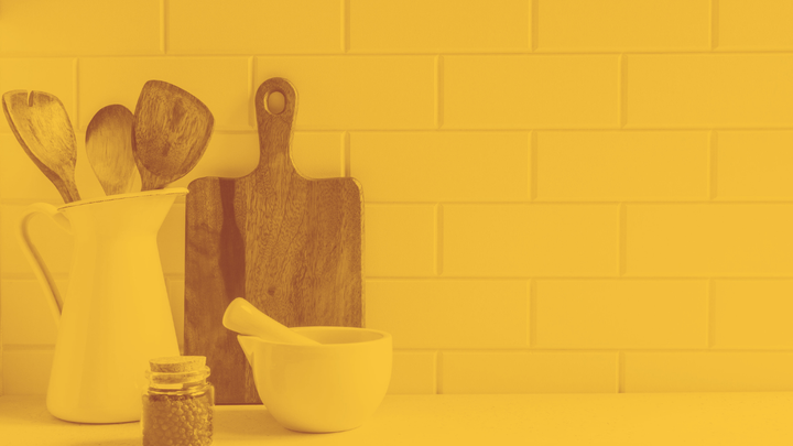 A photo of a cutting board, mortar and pestle, spice jar and pitcher with wooden spoons in it sitting on a kitchen counter.