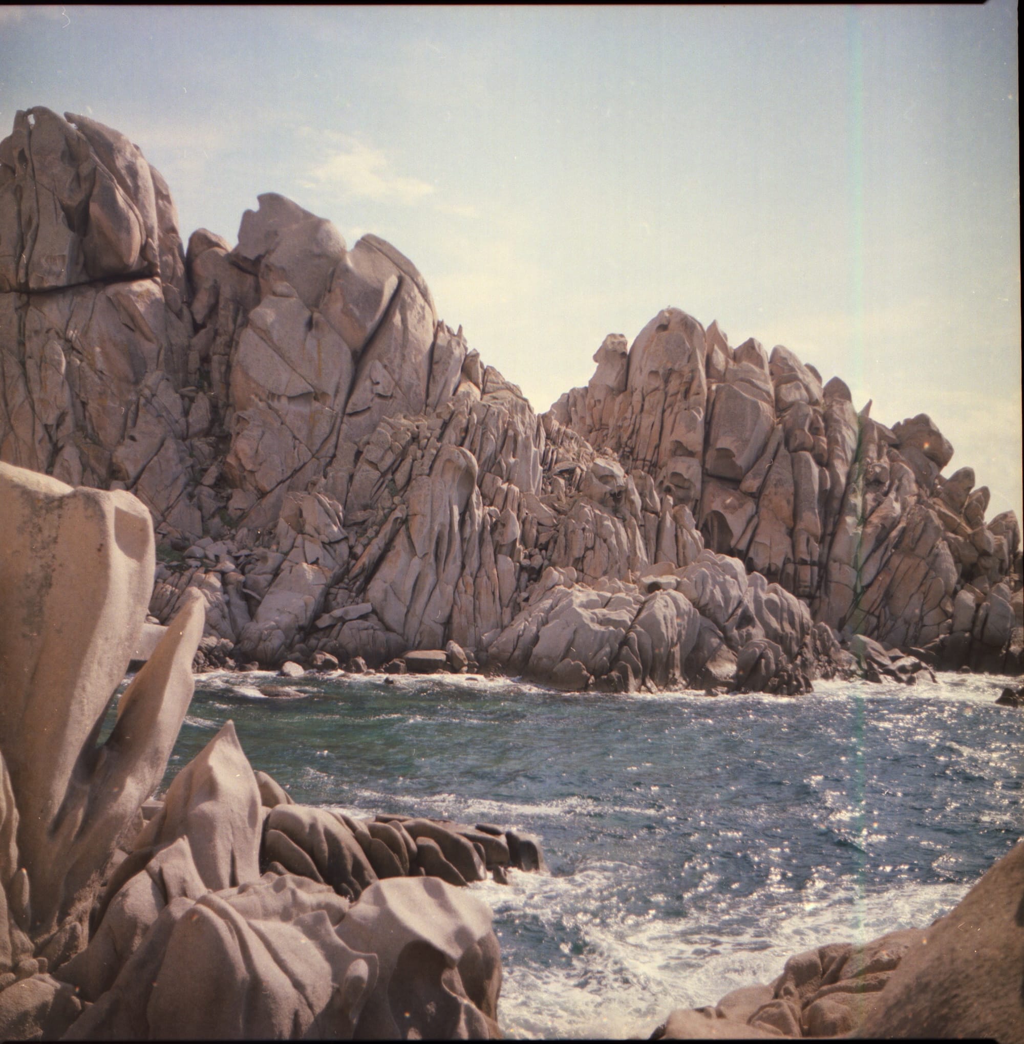 Jagged, weathered rocks jut out from the shoreline, with clear blue water gently lapping against them.