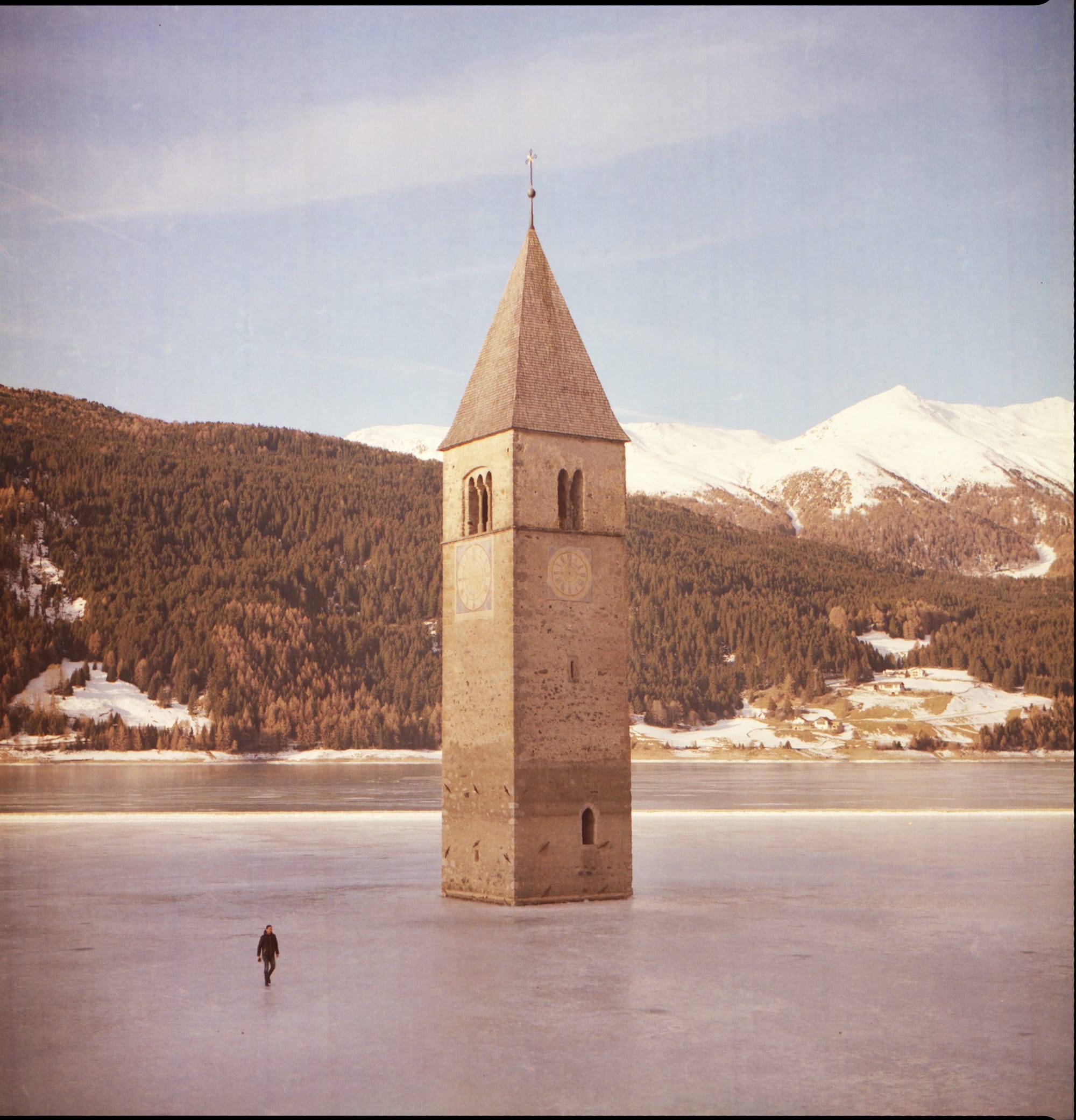 The bell tower of Curon stands in the middle of frozen Lake Resia, surrounded by snow-covered mountains and forests.