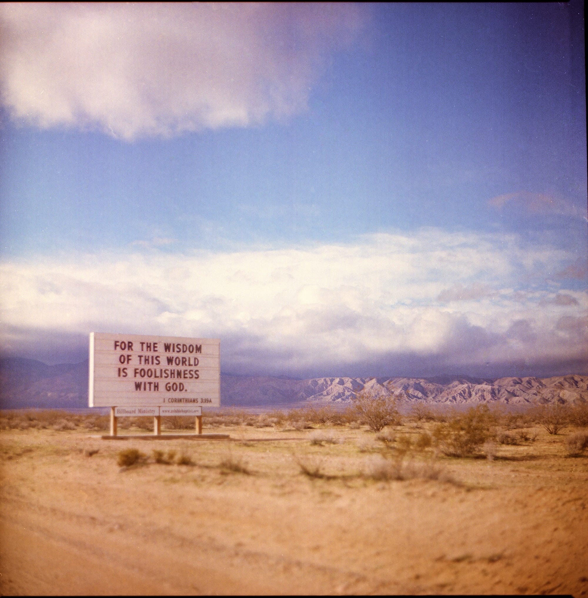 A billboard in a desert landscape with the text 'For the wisdom of this world is foolishness with God. 1 Corinthians 3:19' and a mountain range in the background.
