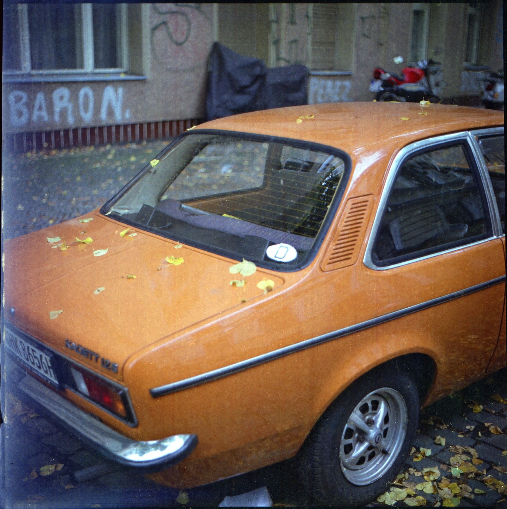 An orange vintage car parked on a leaf-covered street with graffiti on the wall behind it.