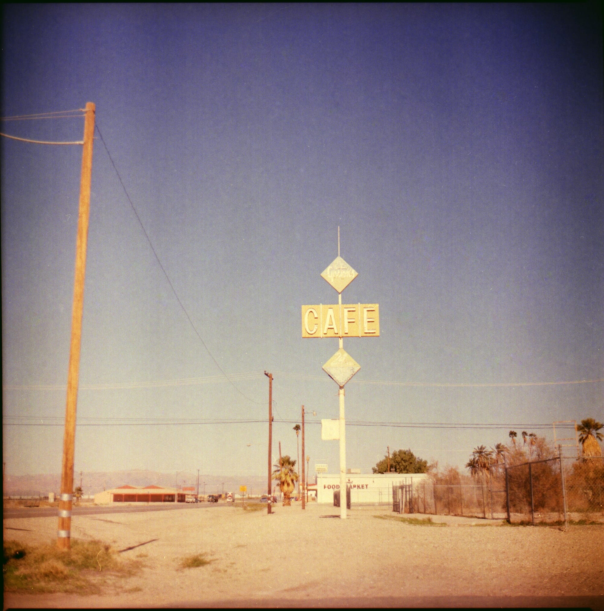 A retro café sign stands tall in a desert-like area with a few scattered buildings and palm trees in the background.