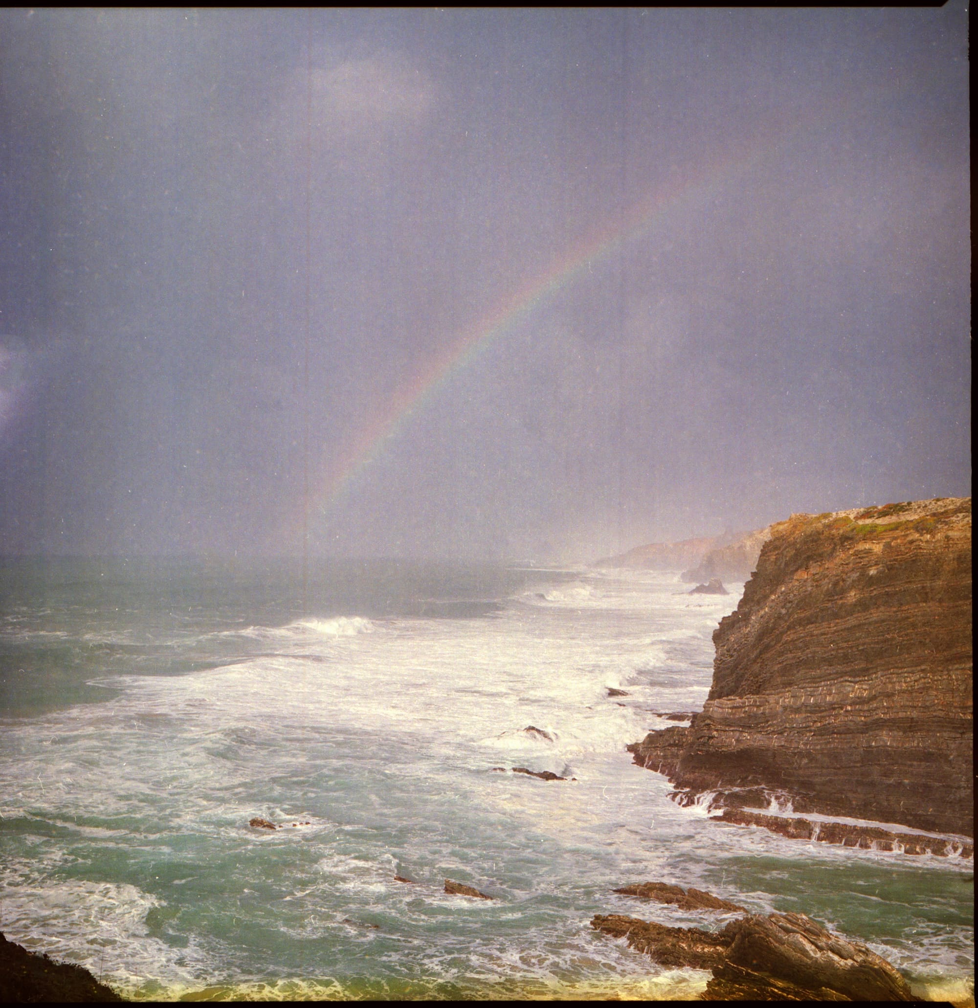 A faint rainbow arches over a stormy sea, with waves crashing against a rocky cliff under a moody sky.
