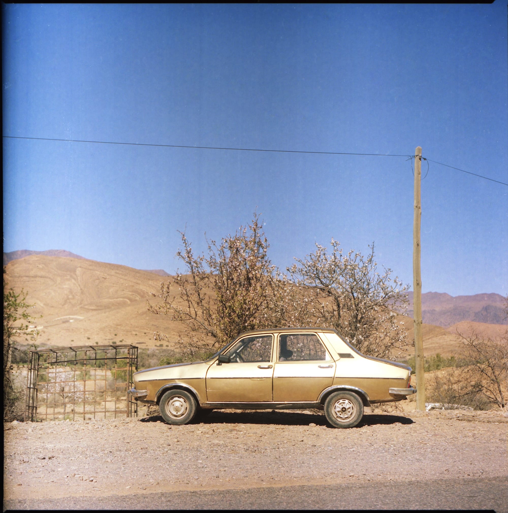 A vintage car parked on a dirt road next, with dry shrubs and a mountainous landscape in the background.