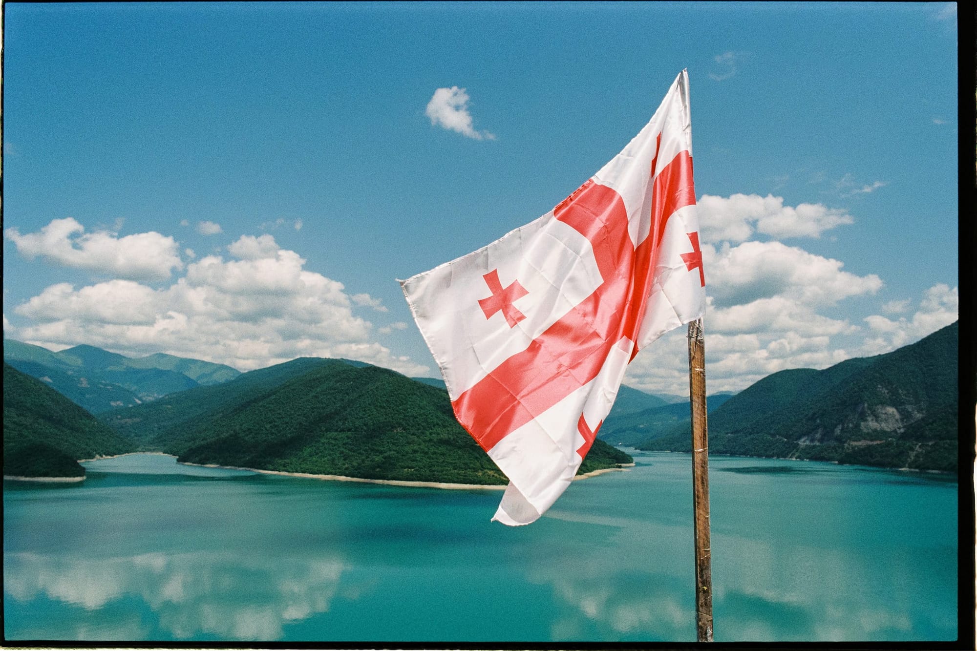 The flag of Georgia flying in a scenic outdoor setting with mountains and a lake.
