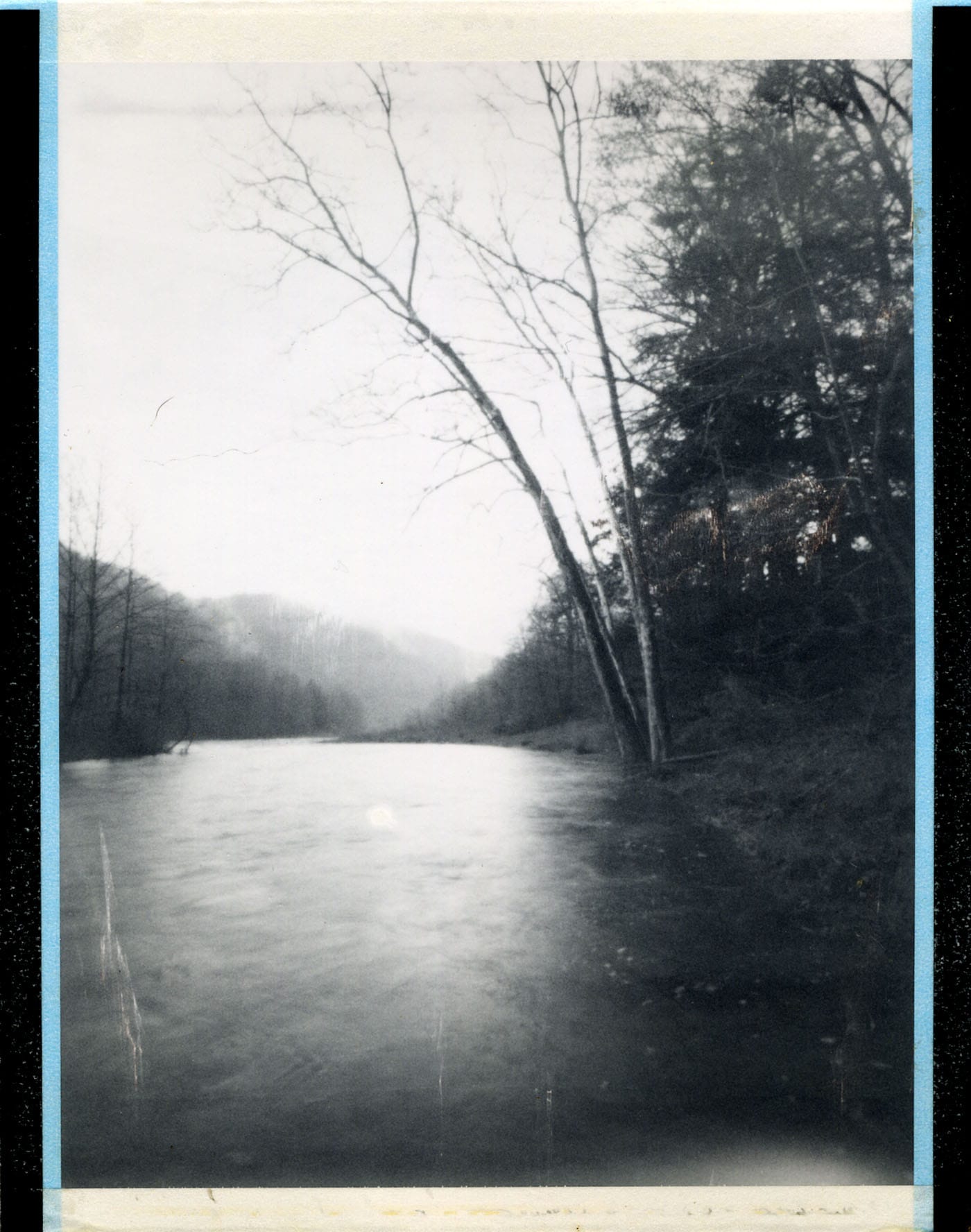 Pinhole black and white photo of a calm river surrounded by leafless trees and foggy woods.