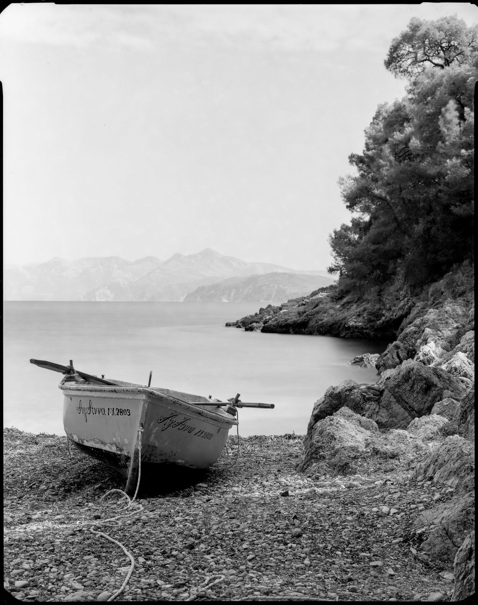 Black and white medium format film photo of a small boat on a rocky shore with a calm sea and distant mountains.