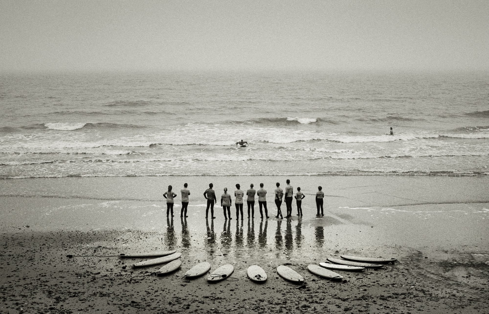 Group of surfers standing on beach with surfboards, ocean waves in the background.