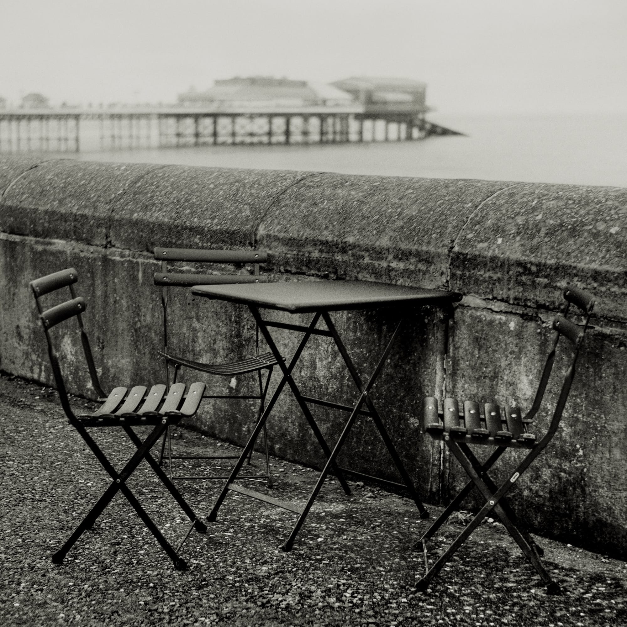 Black and white photo of two empty folding chairs and a table on a stone surface near the sea.